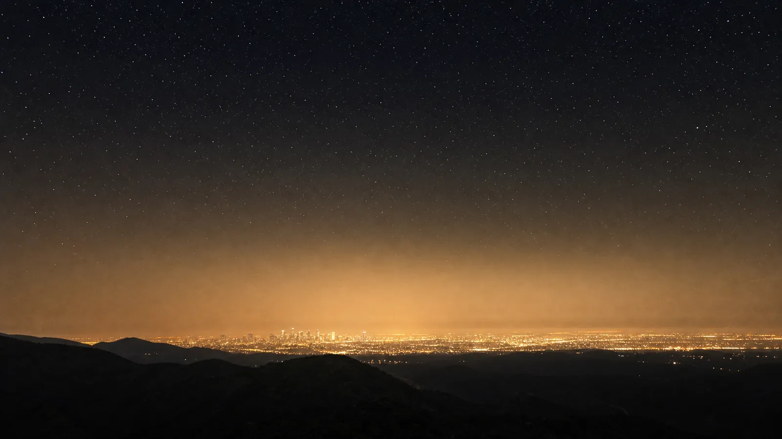 A distant city casts a broad dome of light pollution into a fading star field.