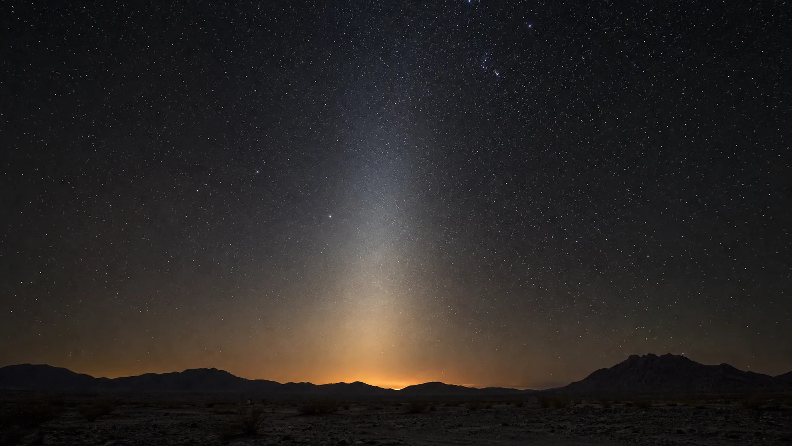 A pale pyramid of zodiacal light rises from a desert horizon into a starry sky.