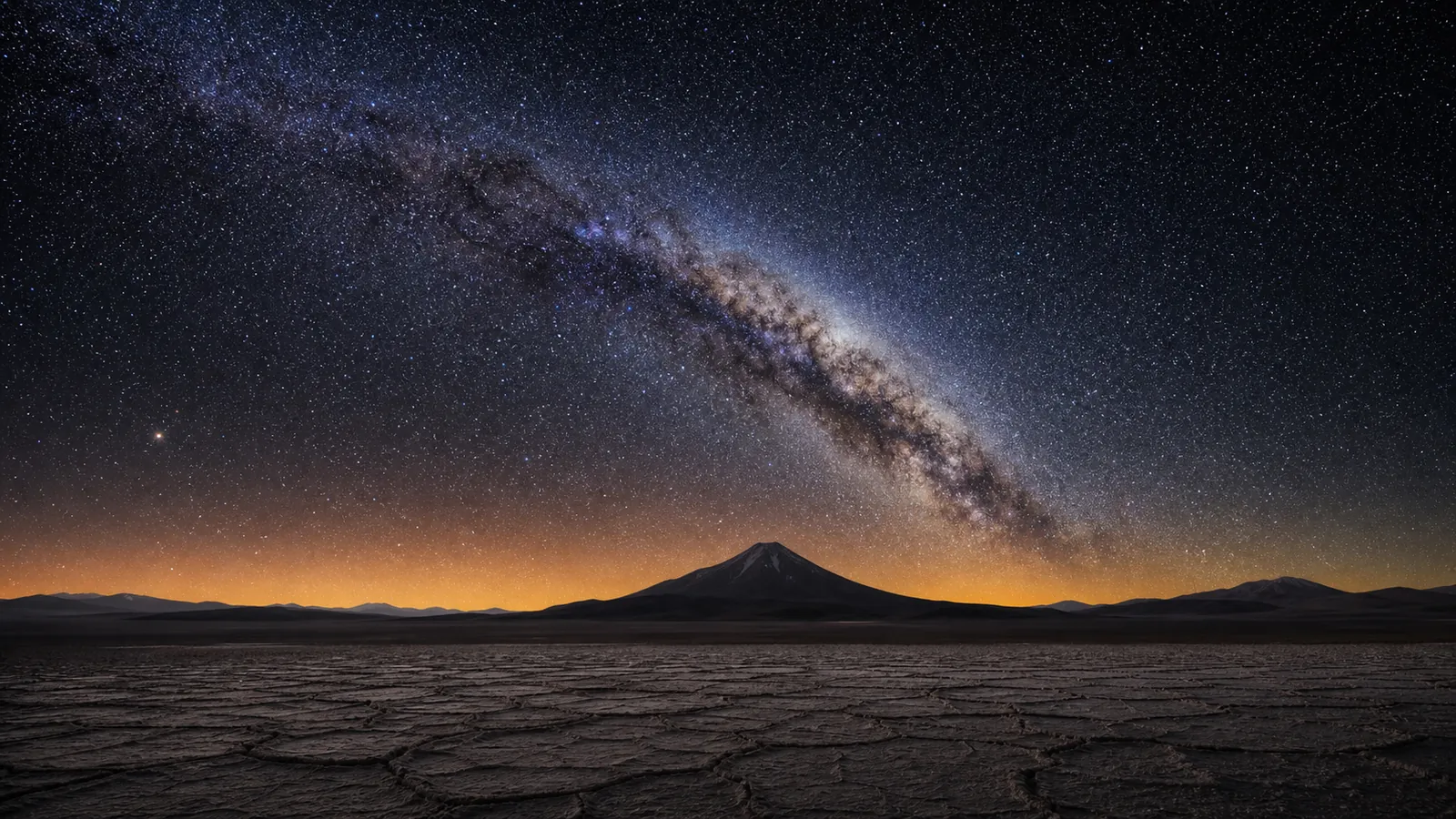 The Milky Way above a cracked Atacama salt flat and distant volcano at night.