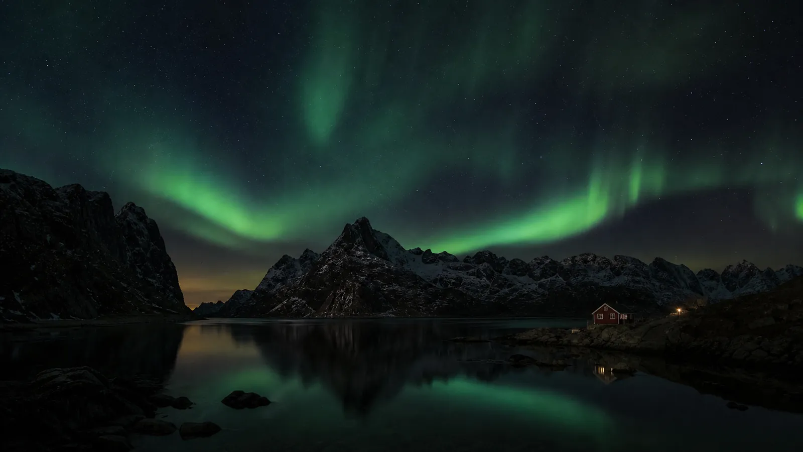 Green aurora over Lofoten's jagged peaks, dark fjord, and a distant red cabin.