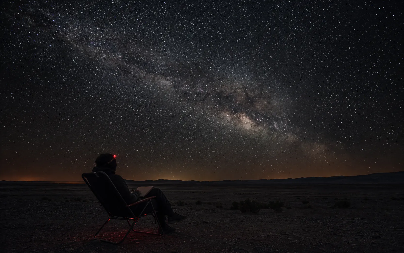 A lone observer reclines with a notebook beneath the Milky Way and a faint red headlamp.